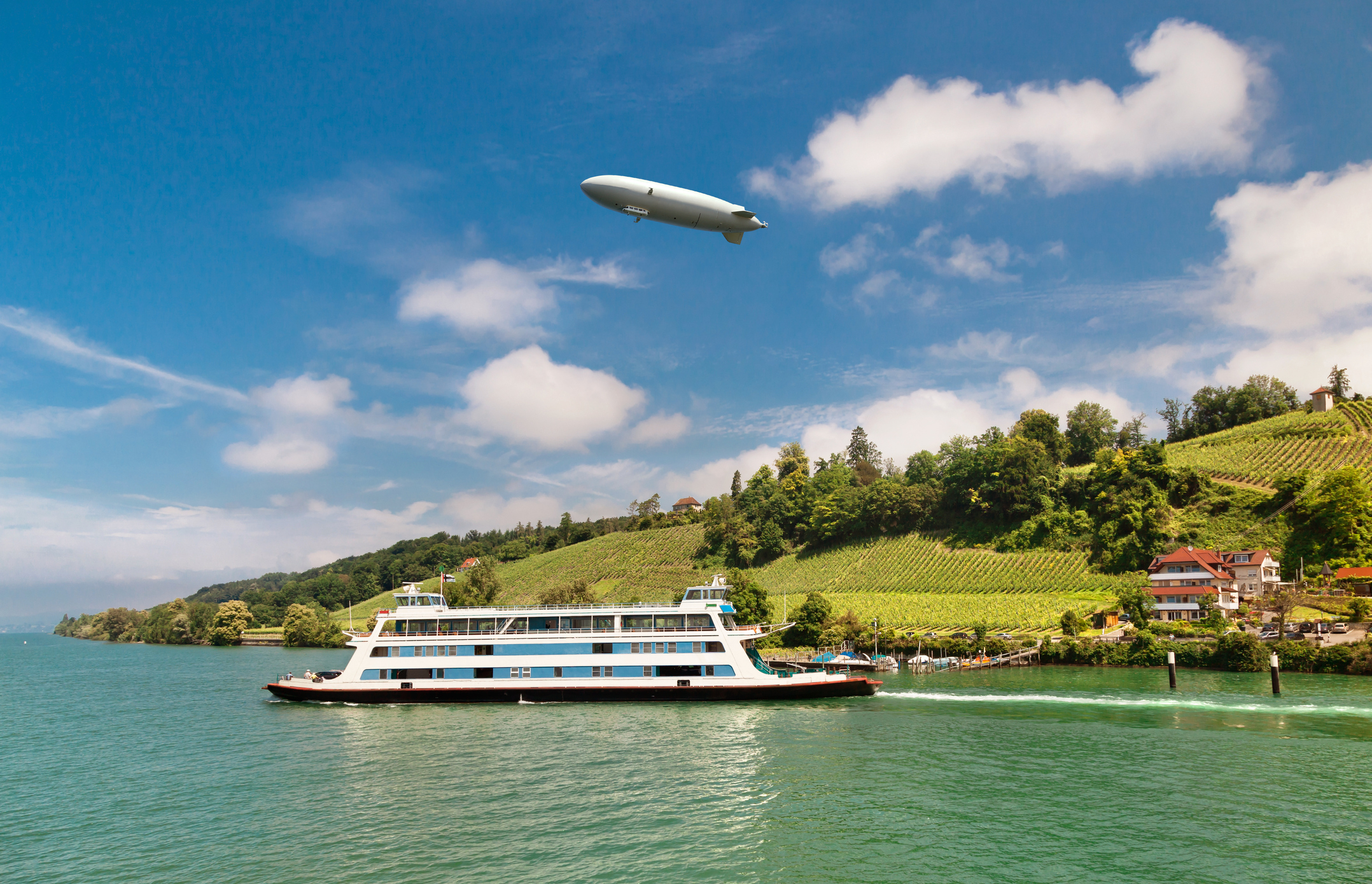 Een Zeppelin zweeft boven een rivierlandschap met wijngaarden en een veerboot.