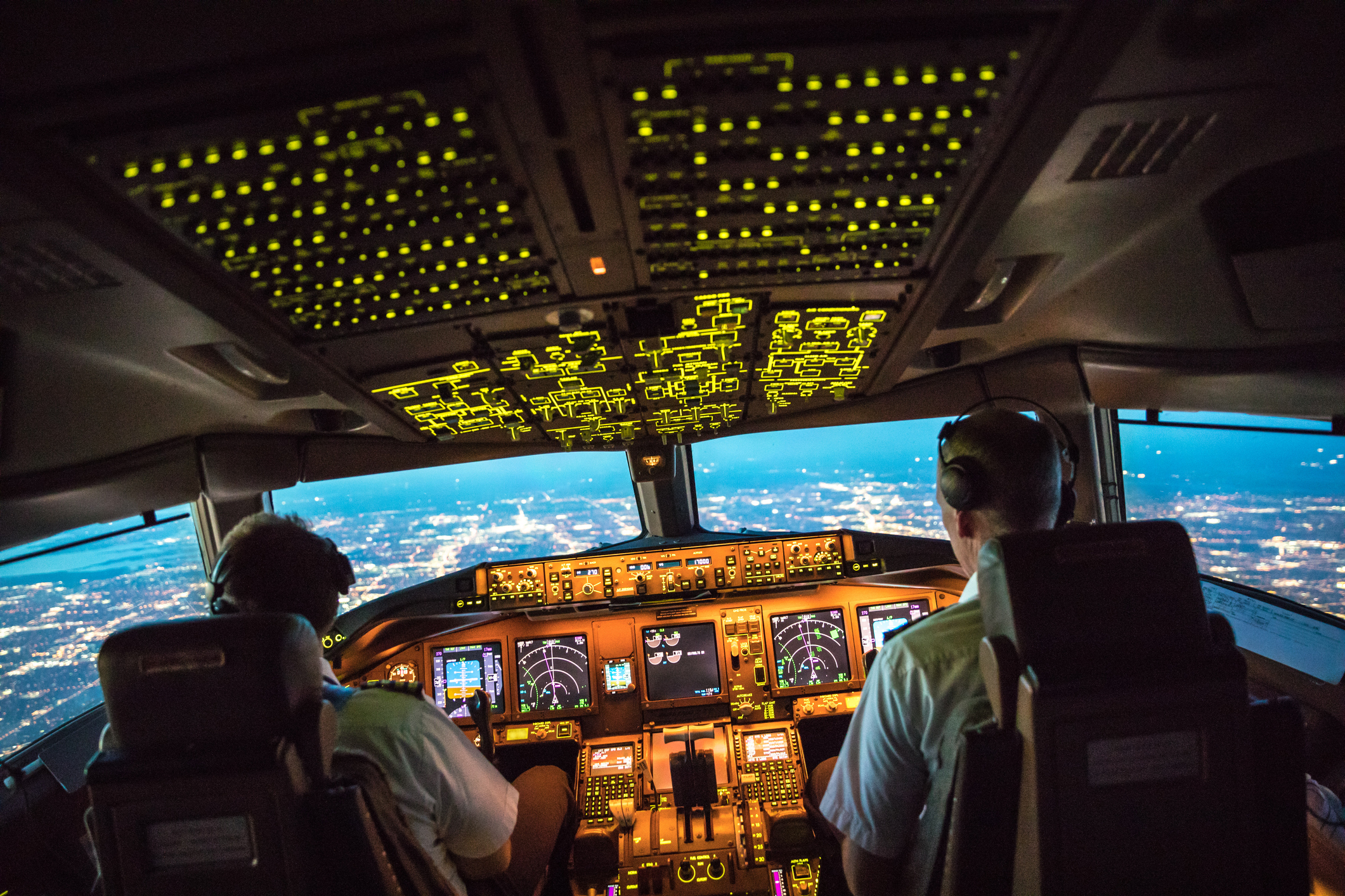 Een verlichte cockpit van een vliegtuig met twee piloten die 's avonds een stad naderen.