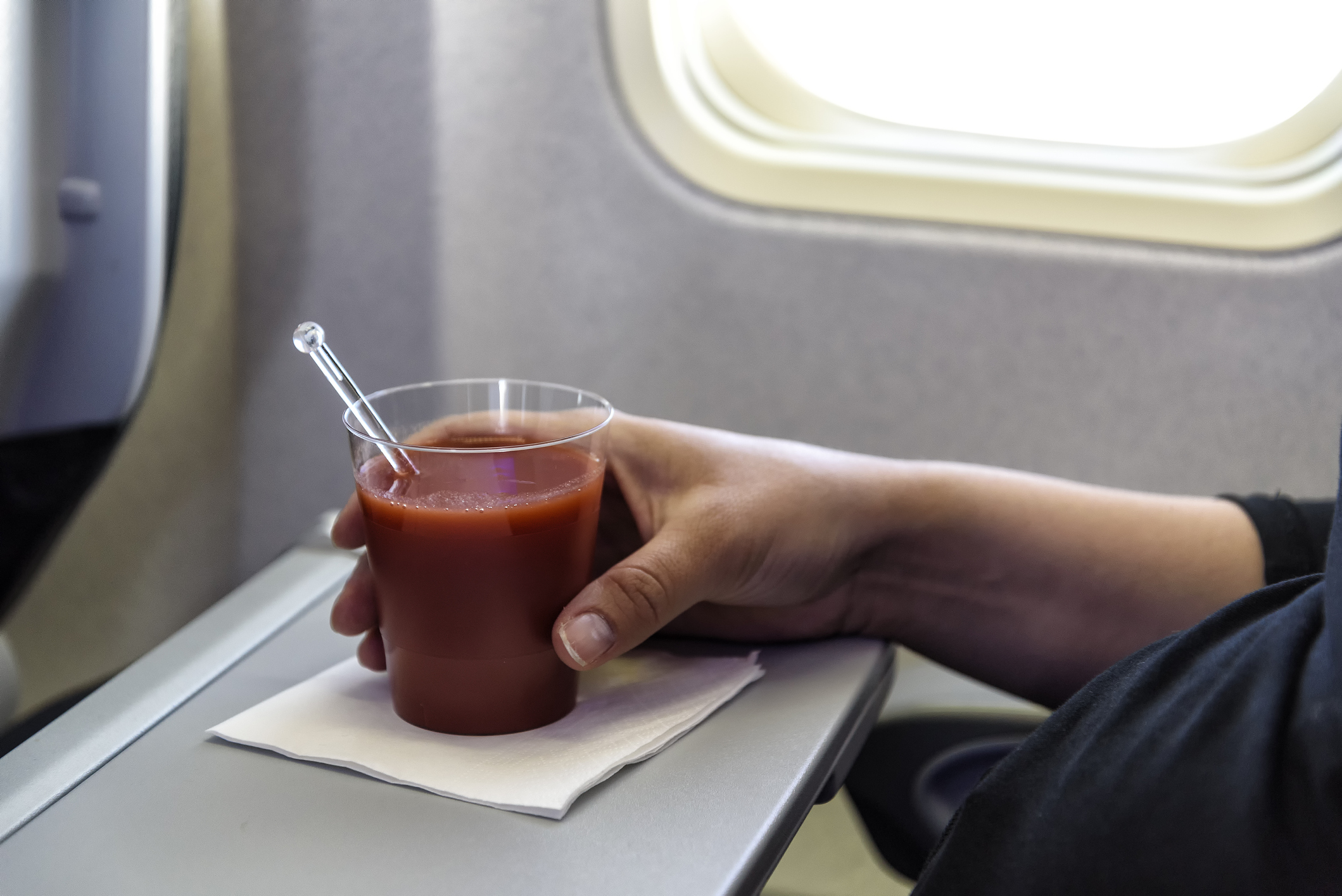Close-up van een hand die een plastic beker tomatensap vastgrijpt die op een servet van een klaptafel in een vliegtuig wordt geplaatst