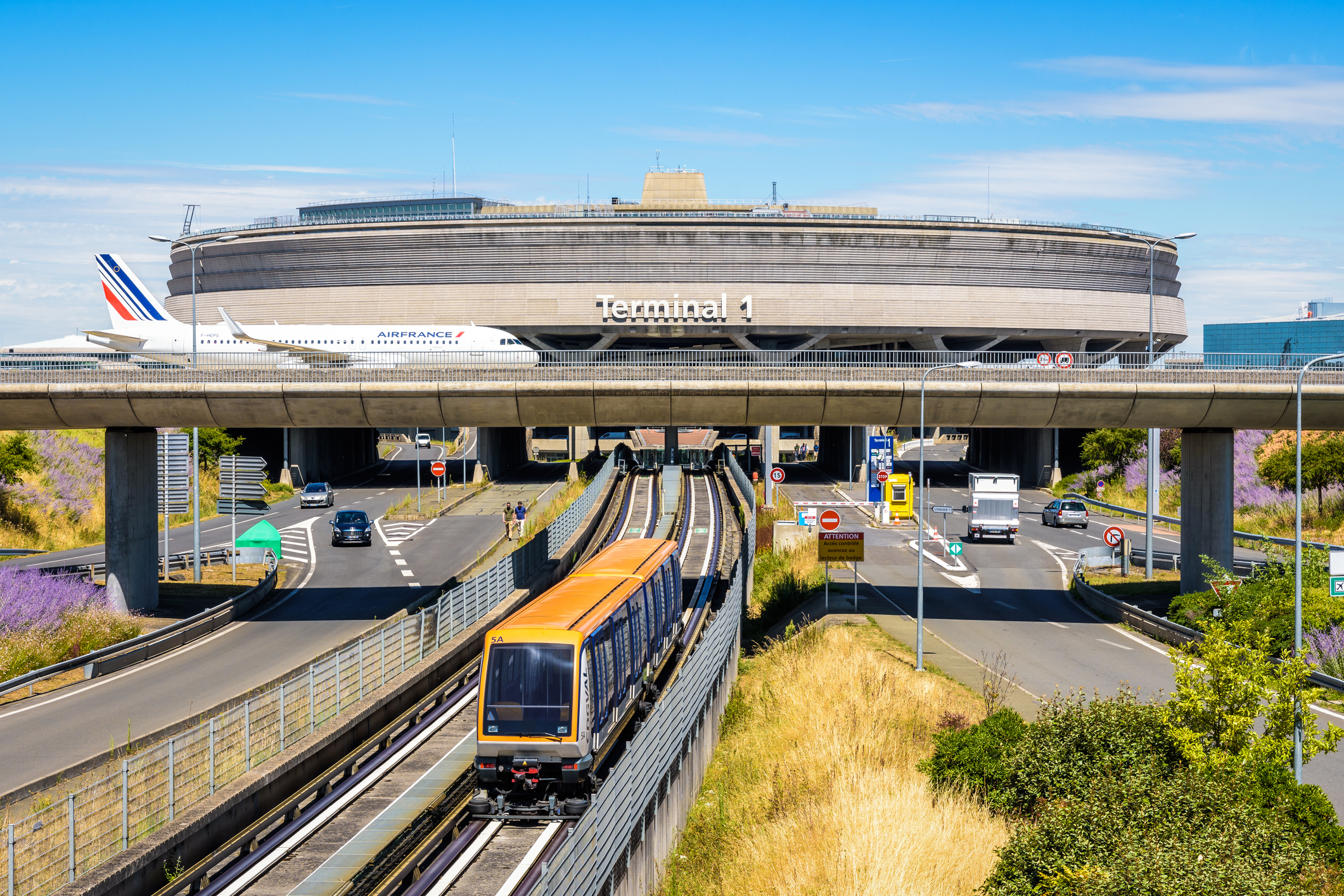 Een afbeelding van Terminal 1 van Charles de Gaulle Airport, op de voorgrond is een tram te zien, op de achtergrond steekt een vliegtuig van Air France op een brug een snelweg over.