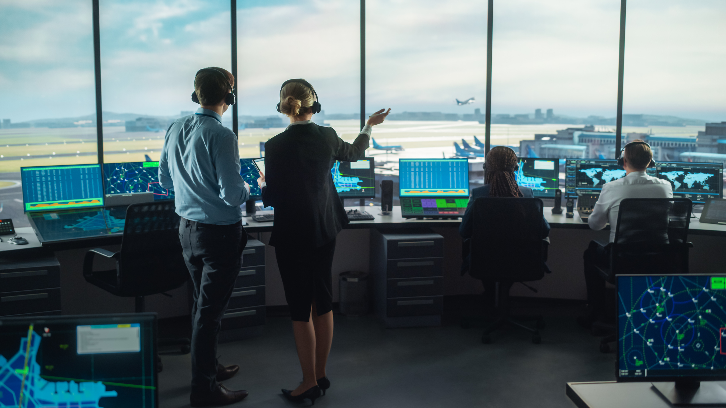 Een luchthaventoren herbergt verschillende luchtverkeersleiders en een paar schermen; start- en landingsbanen en een opstijgend vliegtuig zijn op de achtergrond te zien door de voorruit.