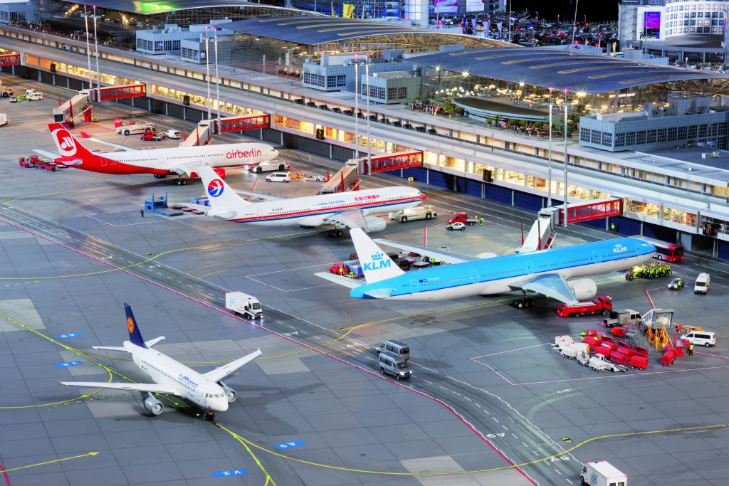 Verschillende vliegtuigen staan geparkeerd met aangedockte gangen aan de gates, op de voorgrond stijgt een Lufthansa vliegtuig op, op de achtergrond zijn de terminals.