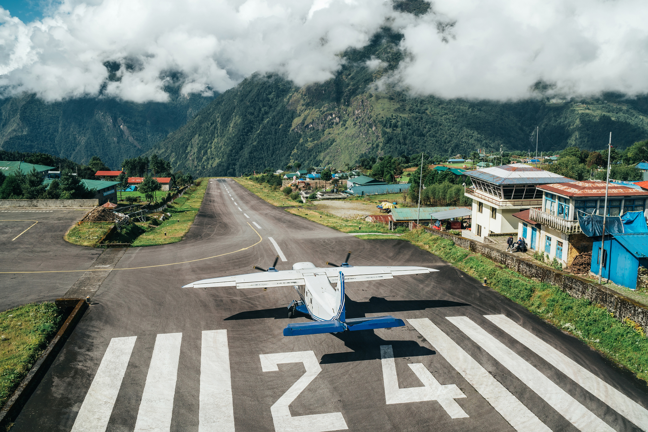 De startbaan in Lukla met zon maar ook enkele laaghangende wolken, een propellervliegtuig begint zojuist met opstijgen.