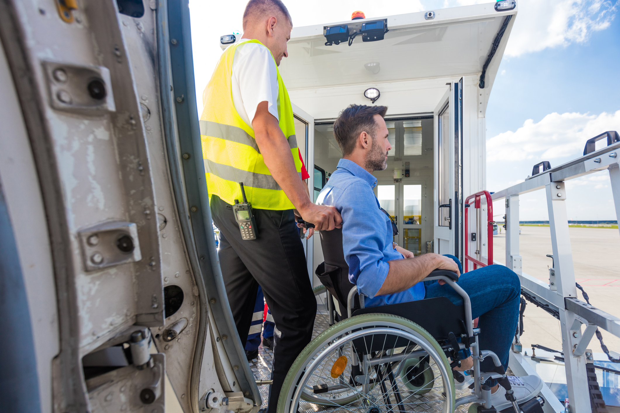Een man in een rolstoel wordt geduwd door een medewerker van een luchtvaartmaatschappij; op de voorgrond is de vliegtuigdeur te zien en op de achtergrond de loopplank.