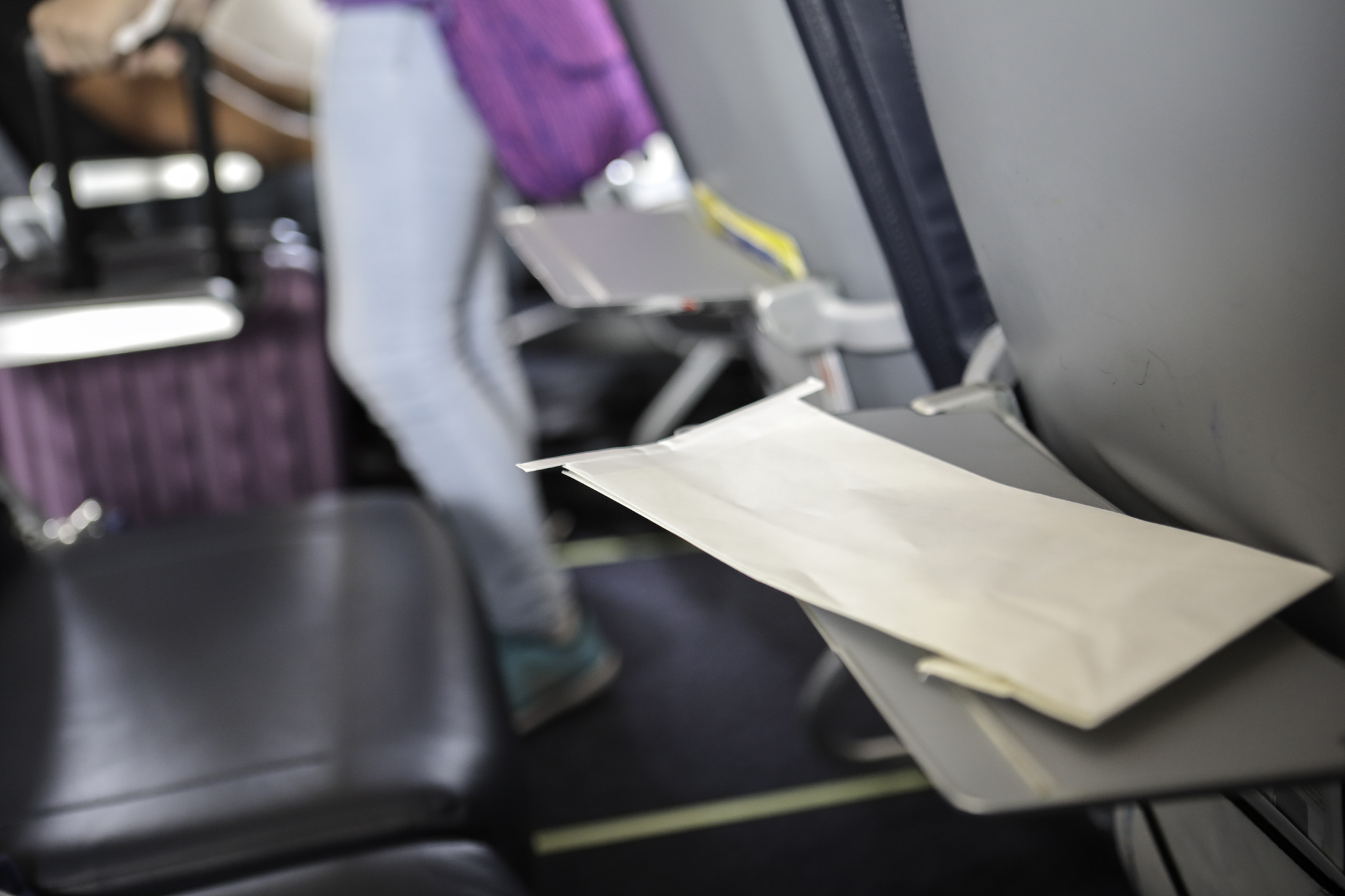 Een enveloppe ligt op een neergeklapt tafeltje bij een vliegtuigstoel, op de achtergrond een andere neergeklapte tafel en een persoon in het gangpad met koffer.