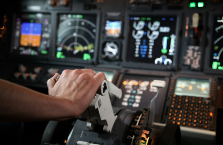 Een hand duwt een bediening naar voren, met op de achtergrond talrijke meters en meters van een cockpit.