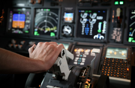 Een hand duwt een bediening naar voren, met op de achtergrond talrijke meters en meters van een cockpit.