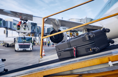 Een koffer wordt met een bagageband in een vliegtuig geladen, op de achtergrond wordt het vliegtuig bijgetankt.