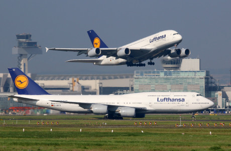 Op de voorgrond staat een Lufthansa vliegtuig op de startbaan van een vliegveld, op de achtergrond stijgt net een tweede vliegtuig op.